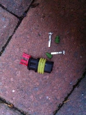 A black two-way connector housing with a yellow seal and red retainer, next to two metal terminals and two green seals, on paving