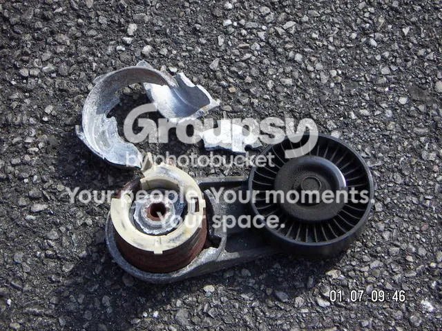 A belt tensioner with its cover in pieces, lying on tarmac next to its pulley