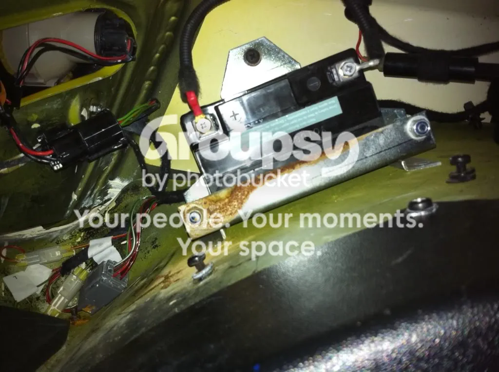 The backup battery in its bracket with the cover removed, a red-banded positive lead on the left terminal and a rust-marked clamp