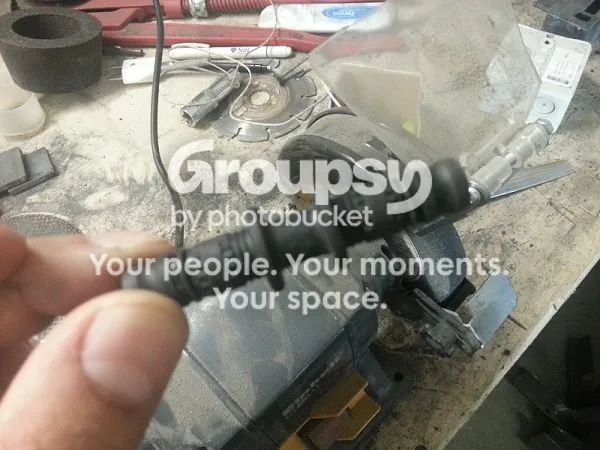 A hand holding a black barbed plastic hose connector in front of a workshop bench