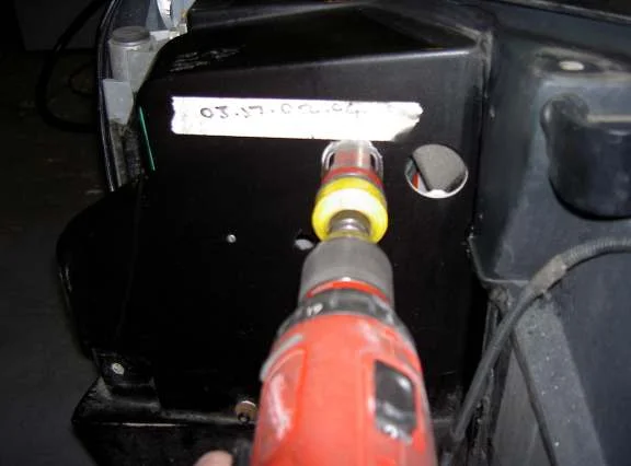 A cordless drill with a hole saw cutting into the black inner sill panel, with a strip of tape marked in pen above