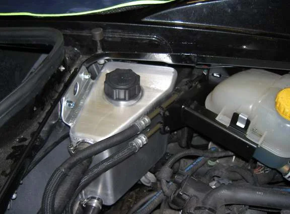 A silver header tank with a black cap beside the original coolant expansion tank in the engine bay