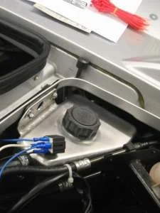 A relay with blue crimped wires attached, beside a silver header tank cap, with red cable ties above