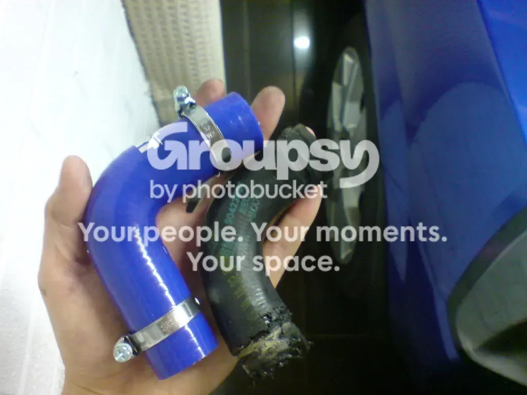A hand holding a blue silicone 90 degree elbow hose beside the old black rubber elbow, whose end is frayed and crumbling