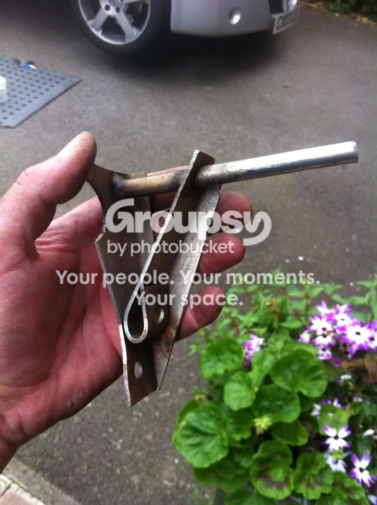A hand holding the failed exhaust hanger bracket: a thin folded steel plate with a tube welded across it and a spring clip, with a sheared edge at the top