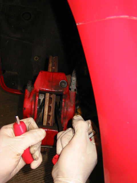 Two gloved hands using small tools inside a red front caliper to push the pistons back, with the disc visible