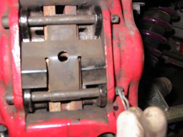The front caliper with two rods in place through the pads, the retaining plate with a round hole between them, and a clip being fitted at the bottom