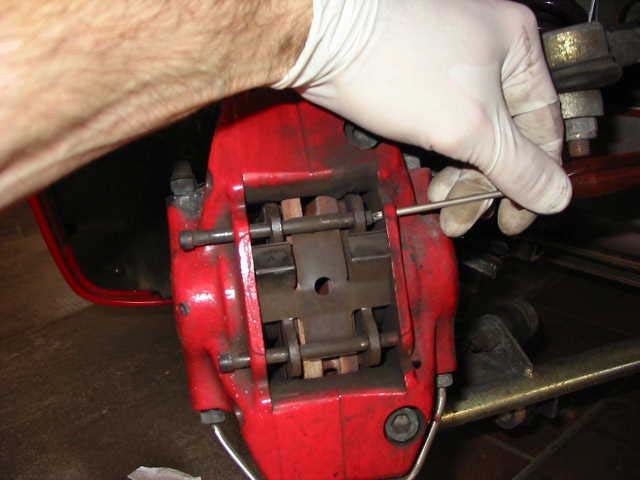 A gloved hand holding a punch against the end of a rod in a red front caliper, with the retaining plate and pads visible