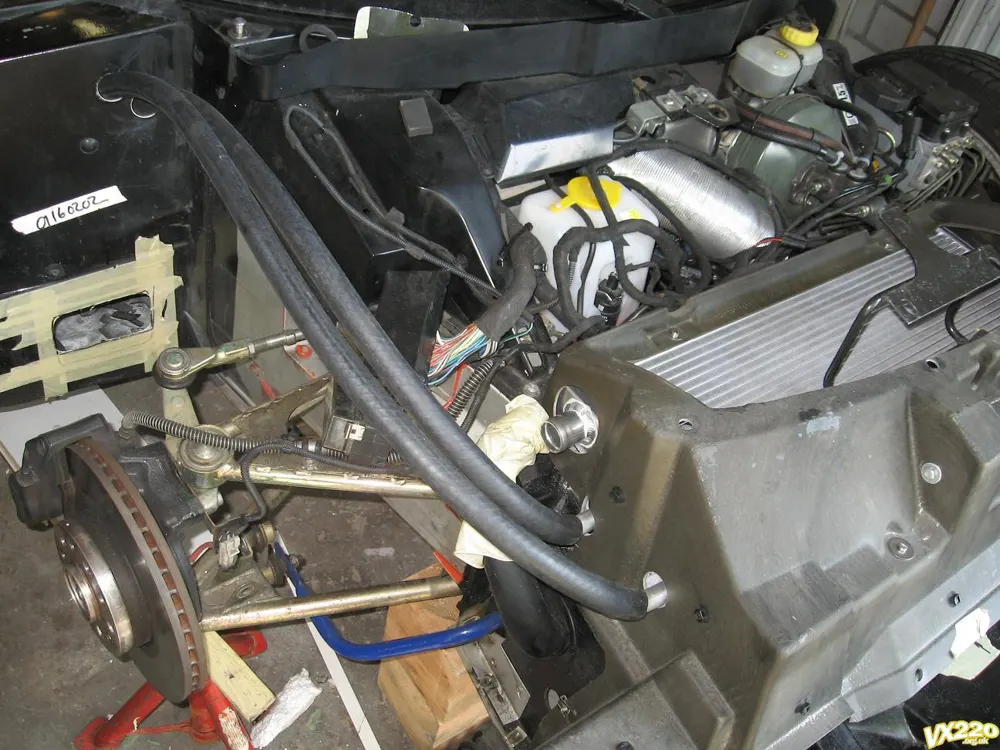 A car with the front clam off, with two black hoses running from the wheel arch area forward to the pre-rad pipes on the side of the radiator housing
