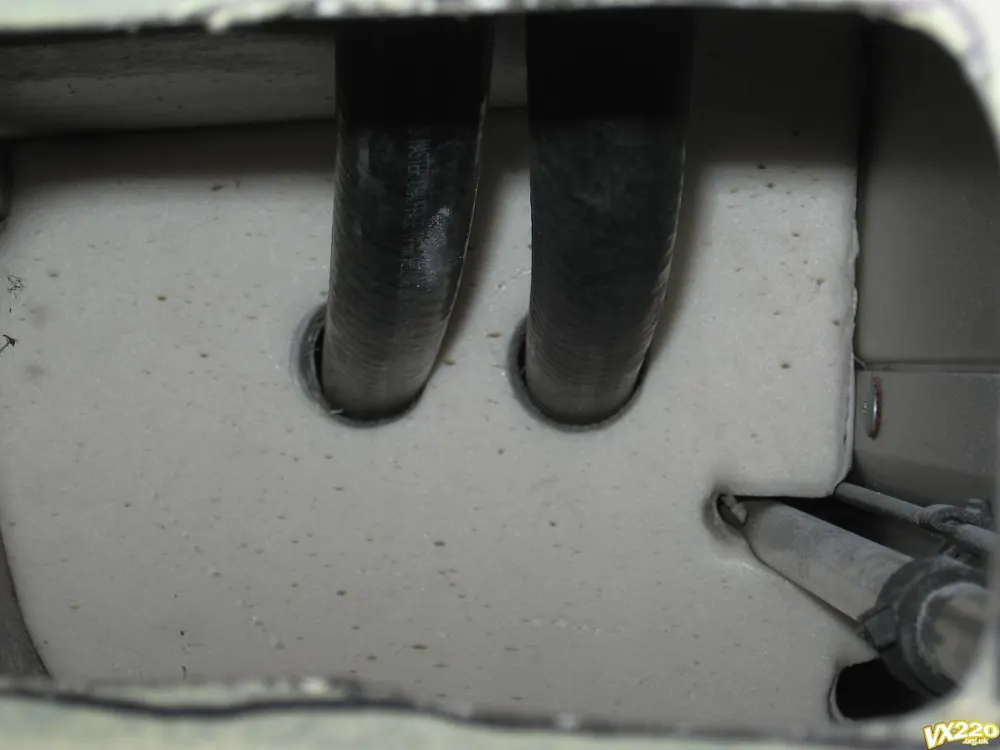 Two black hoses passing through round holes in a block of grey foam inside the sill, with a pipe to the right