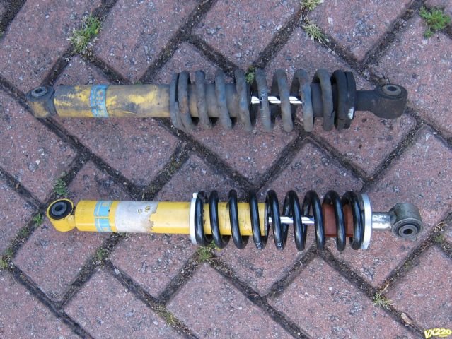 Two yellow shock absorbers lying on block paving, the upper one with a corroded dark spring and the lower one with a black spring and a white spring seat