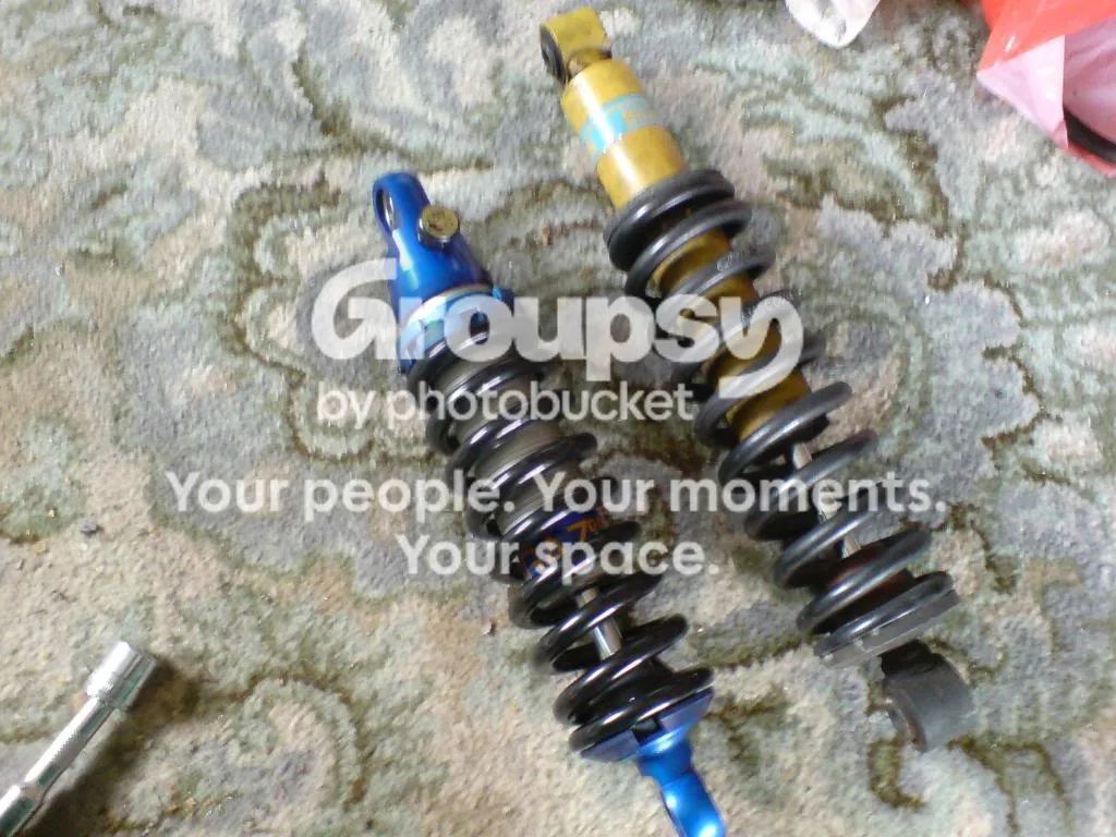 An old yellow Bilstein shock and a new blue Gaz shock with black springs lying on a patterned carpet