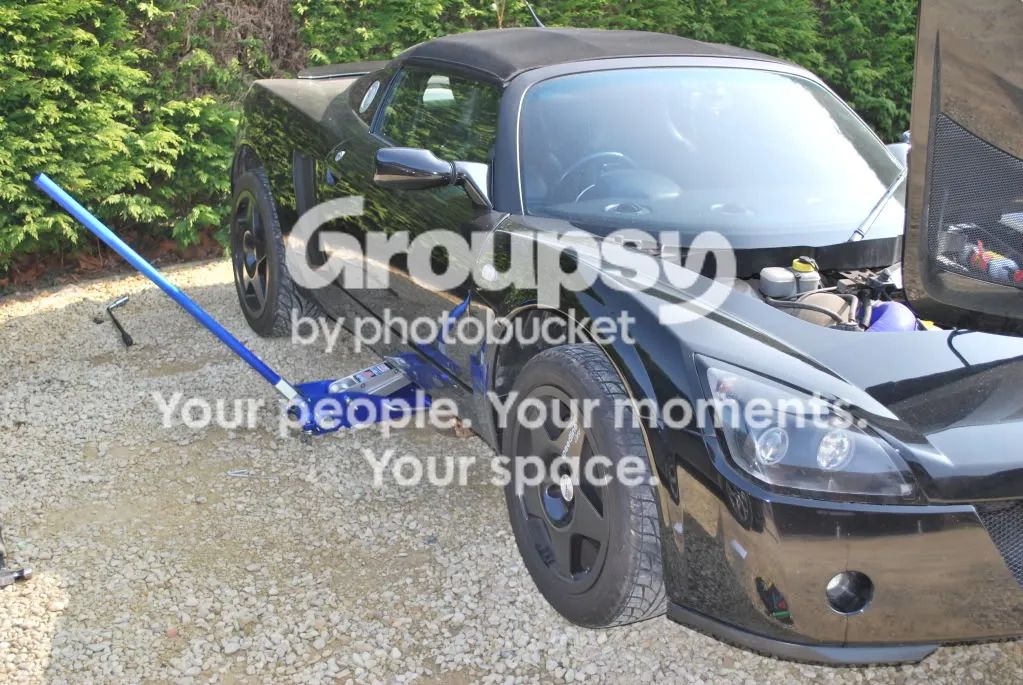 A black car jacked up at the front on gravel, with a blue trolley jack and the bonnet open