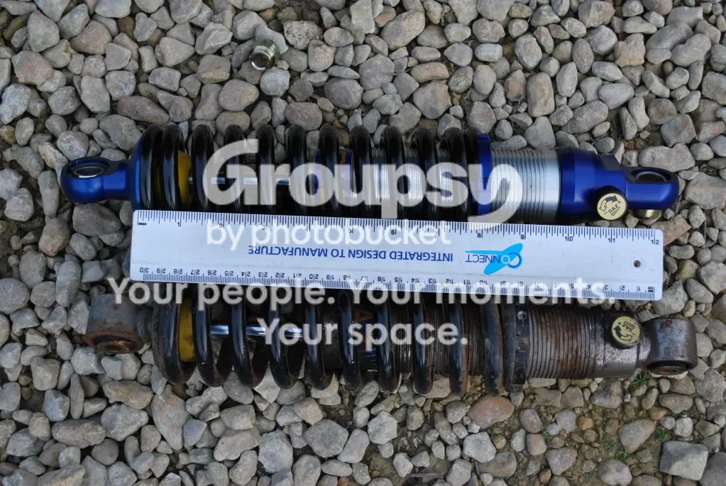 A new blue adjustable shock and an old standard shock lying on gravel next to a ruler