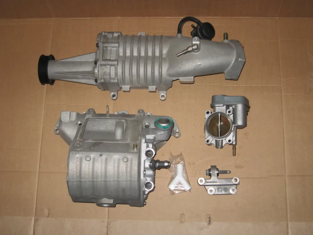 A supercharger with its pulley, a cast inlet manifold, a throttle body, a small cast bracket marked GM and a white plastic Y piece laid out on cardboard