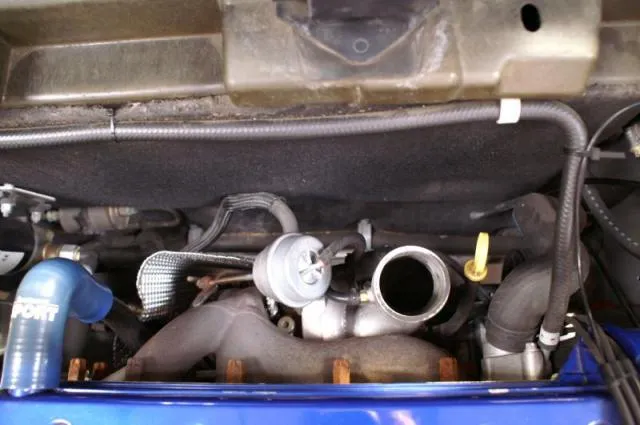 Top view into the engine bay of a blue car with the upper heat shield removed, showing the turbo, a blue silicone hose and the exhaust manifold