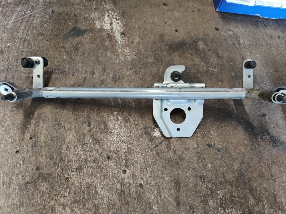 A metal wiper linkage bar with three pivot points and a plate for the motor, on a concrete floor