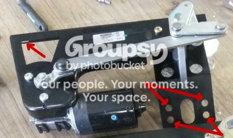 A black wiper motor and linkage plate on a bench, with red arrows drawn on the bolt positions