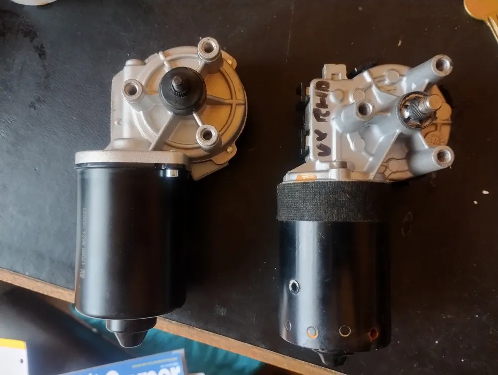 Two wiper motors side by side on a workbench, one silver with a black can and one with a plastic housing